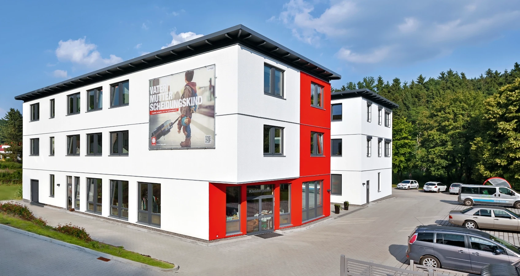 Modernes Gebäude mit weißen Fassaden, roten Akzenten und einem grossen Werbeplakat, Autos auf dem Parkplatz.