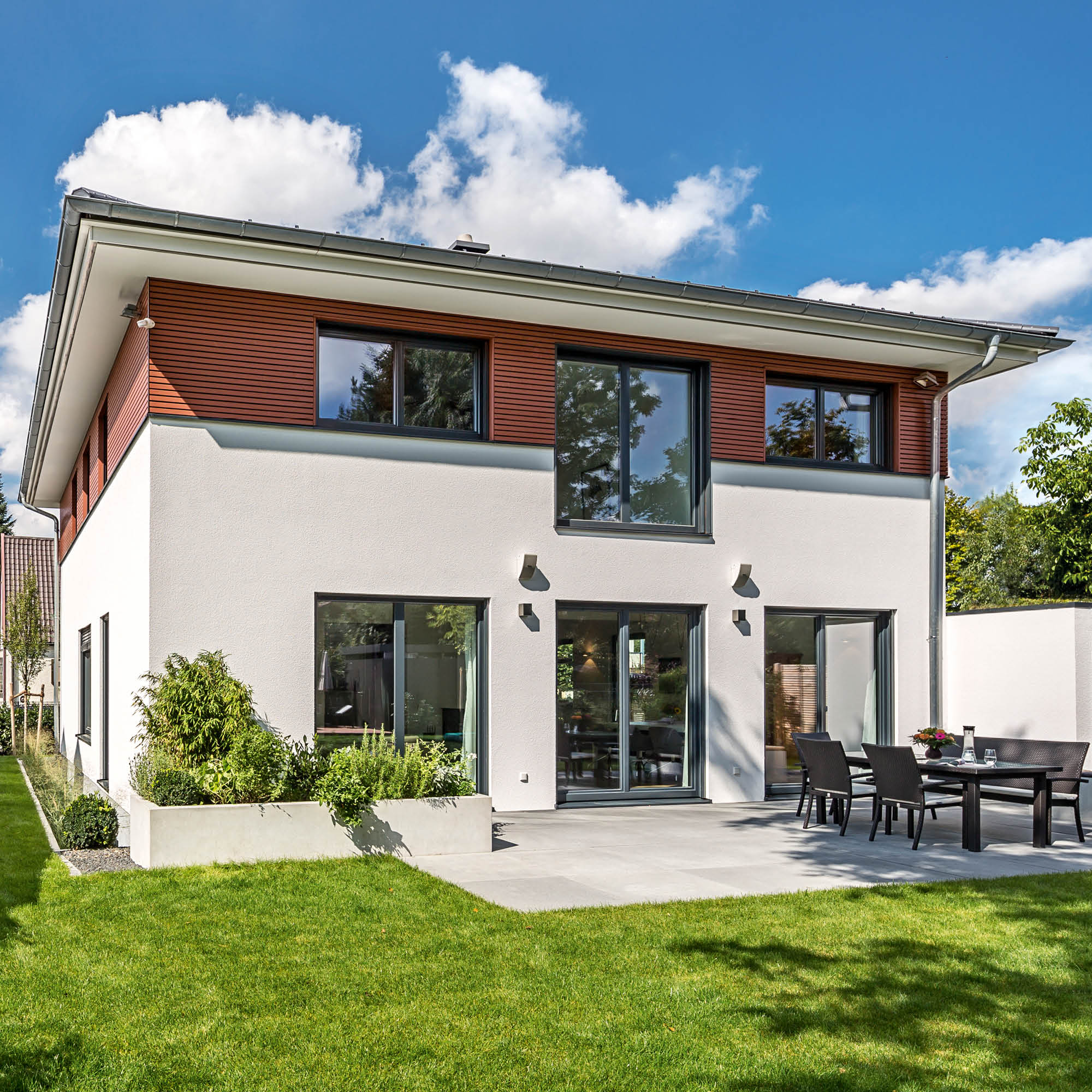 Modernes Haus mit heller Putzfassade, Holzakzenten, großen Fenstern und angeschlossener Terrasse mit Sitzmöbeln.