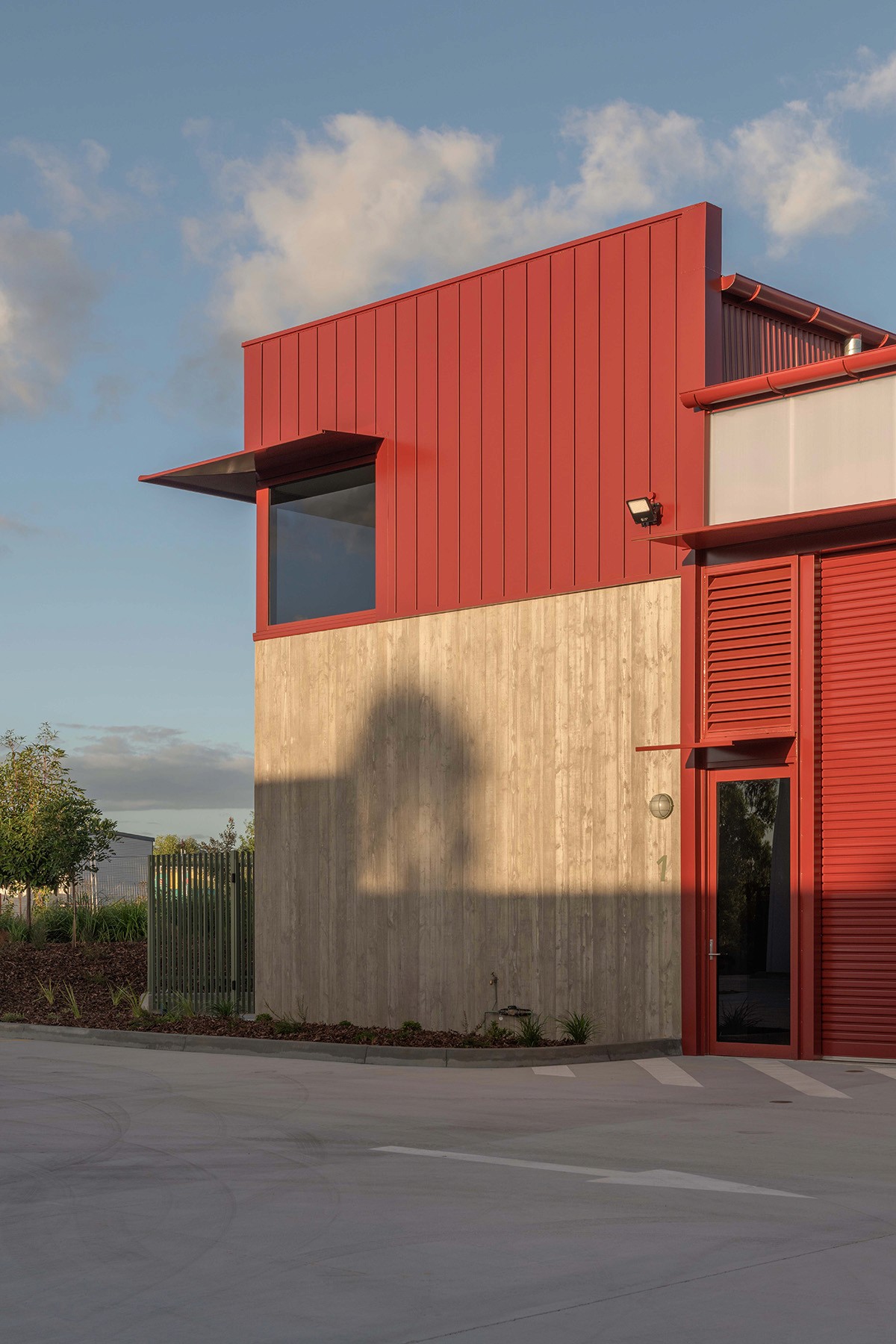 RedSheds exterior detail showing a red metal-clad upper volume above a raw concrete base, with a projecting window canopy and roller door entry set within an open industrial yard.