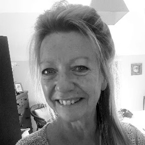 A black and white portrait photo of Belinda Colston. She is smiling and has shoulder-length hair, taken indoors with home furnishings visible in the background.