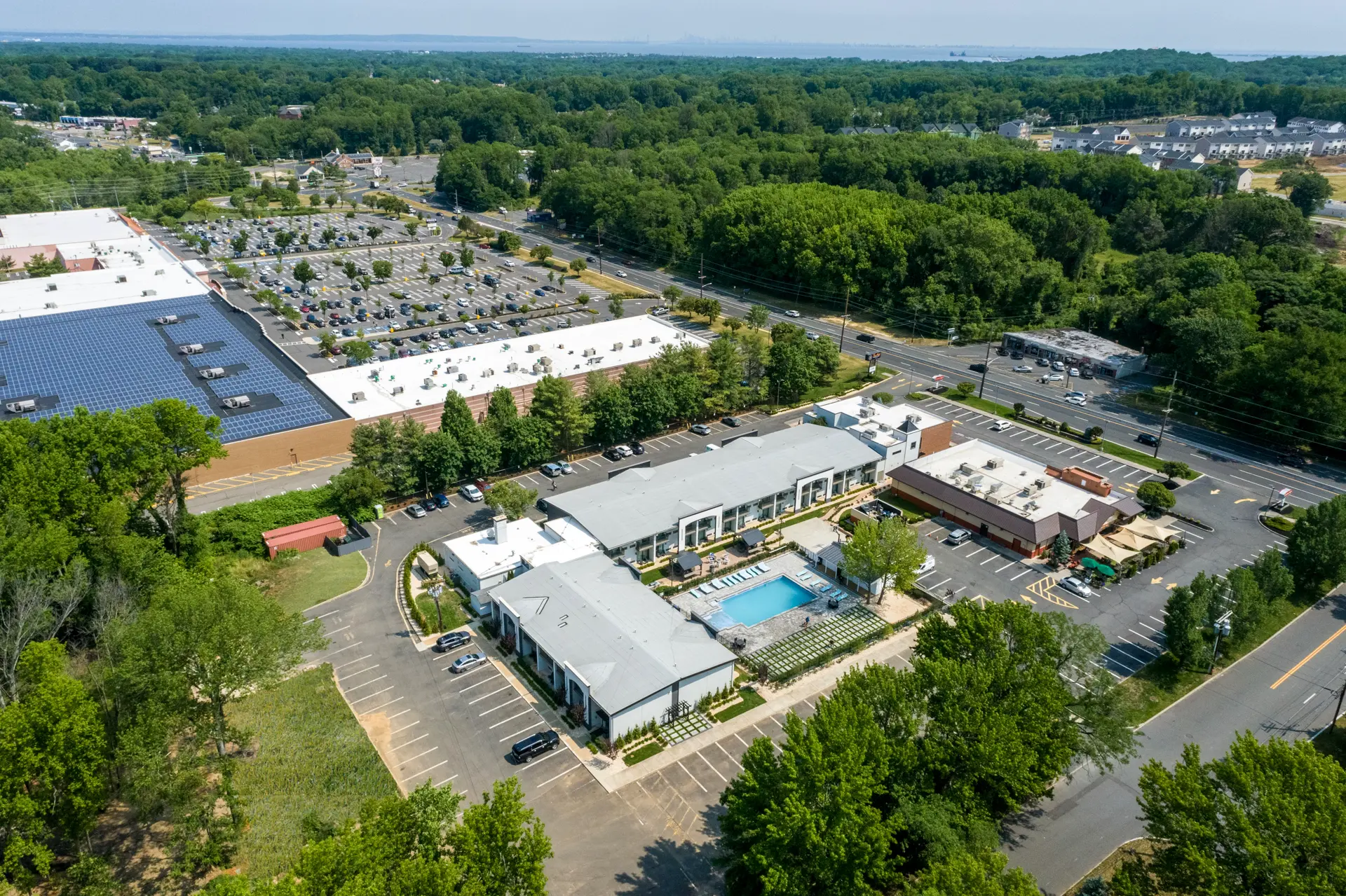 Aerial view of a commercial and residential complex with buildings, parking lots, and a swimming pool surrounded by trees.