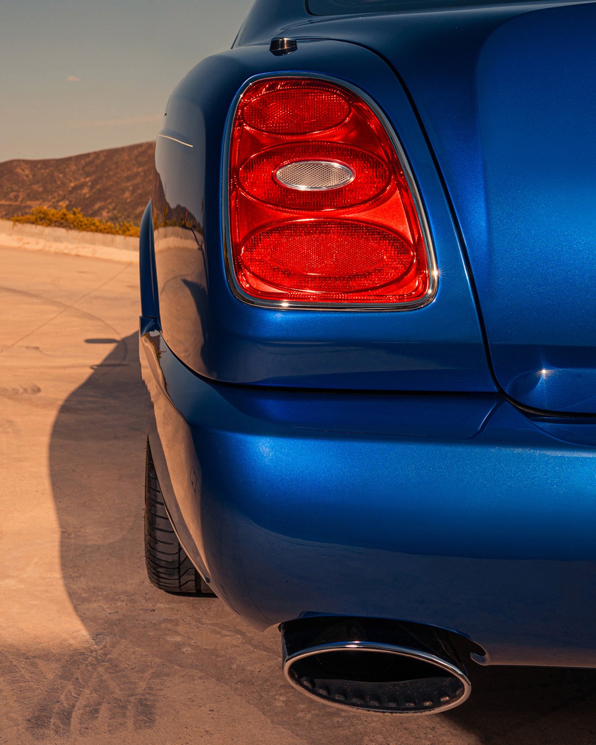 Close-up detail of a Bentley Brooklands rear taillight, the distinctive triple-element red lamp framed in chrome against Moroccan Blue bodywork, polished oval exhaust tip below.