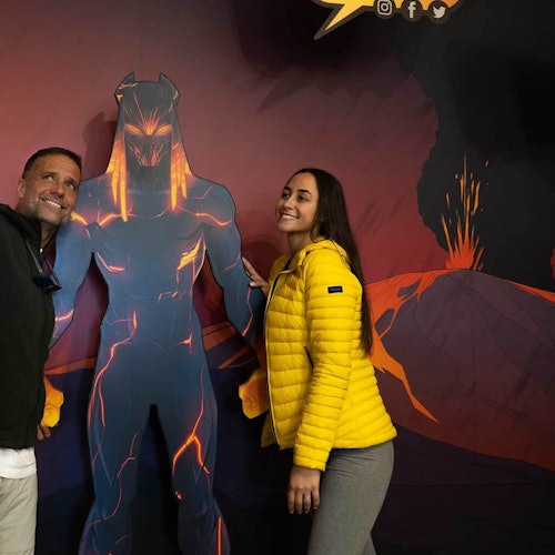 Two people pose next to a cutout of a lava-themed superhero. The background features erupting volcanoes.