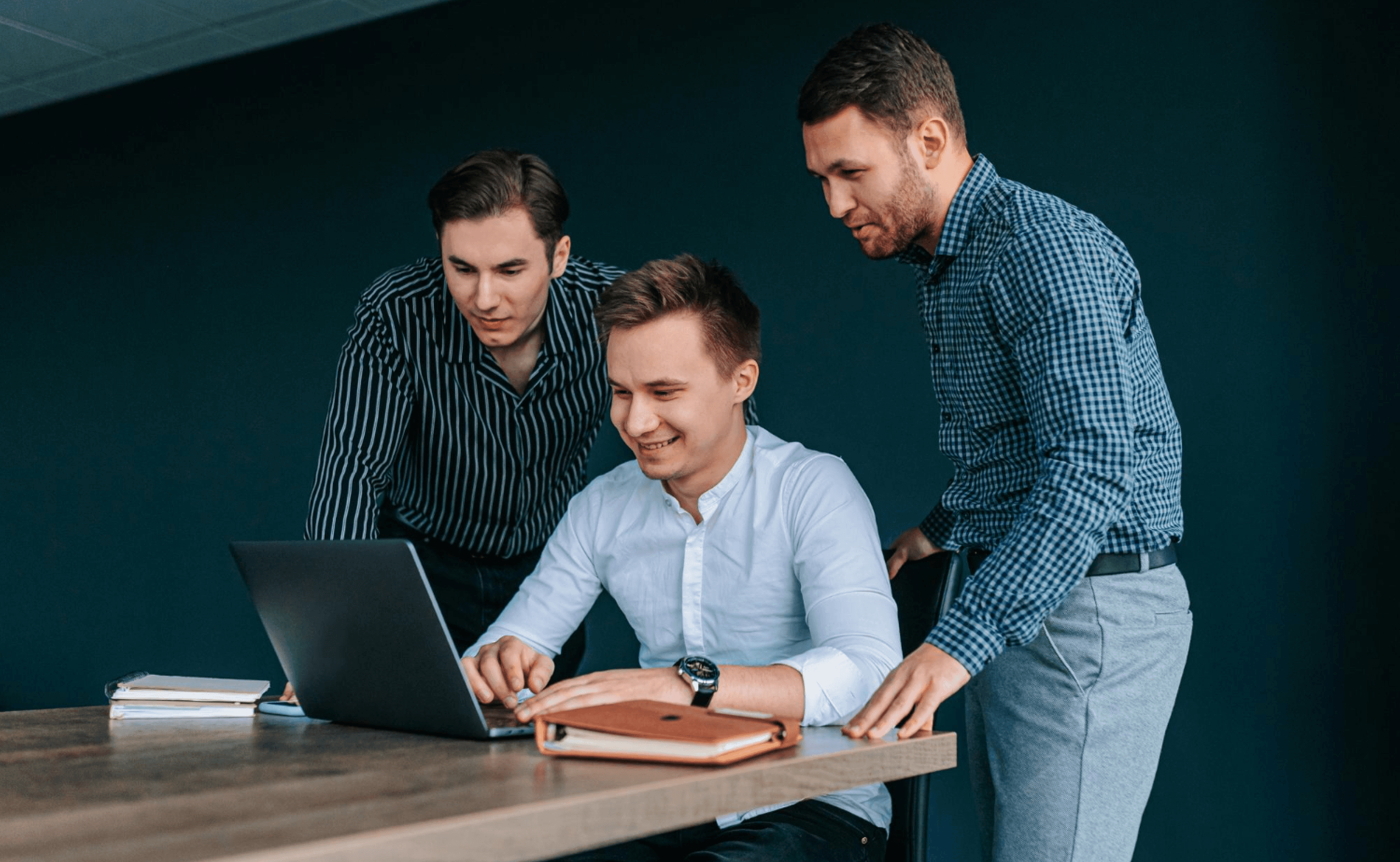 Three people gathered around a laptop, smiling and discussing in a modern office