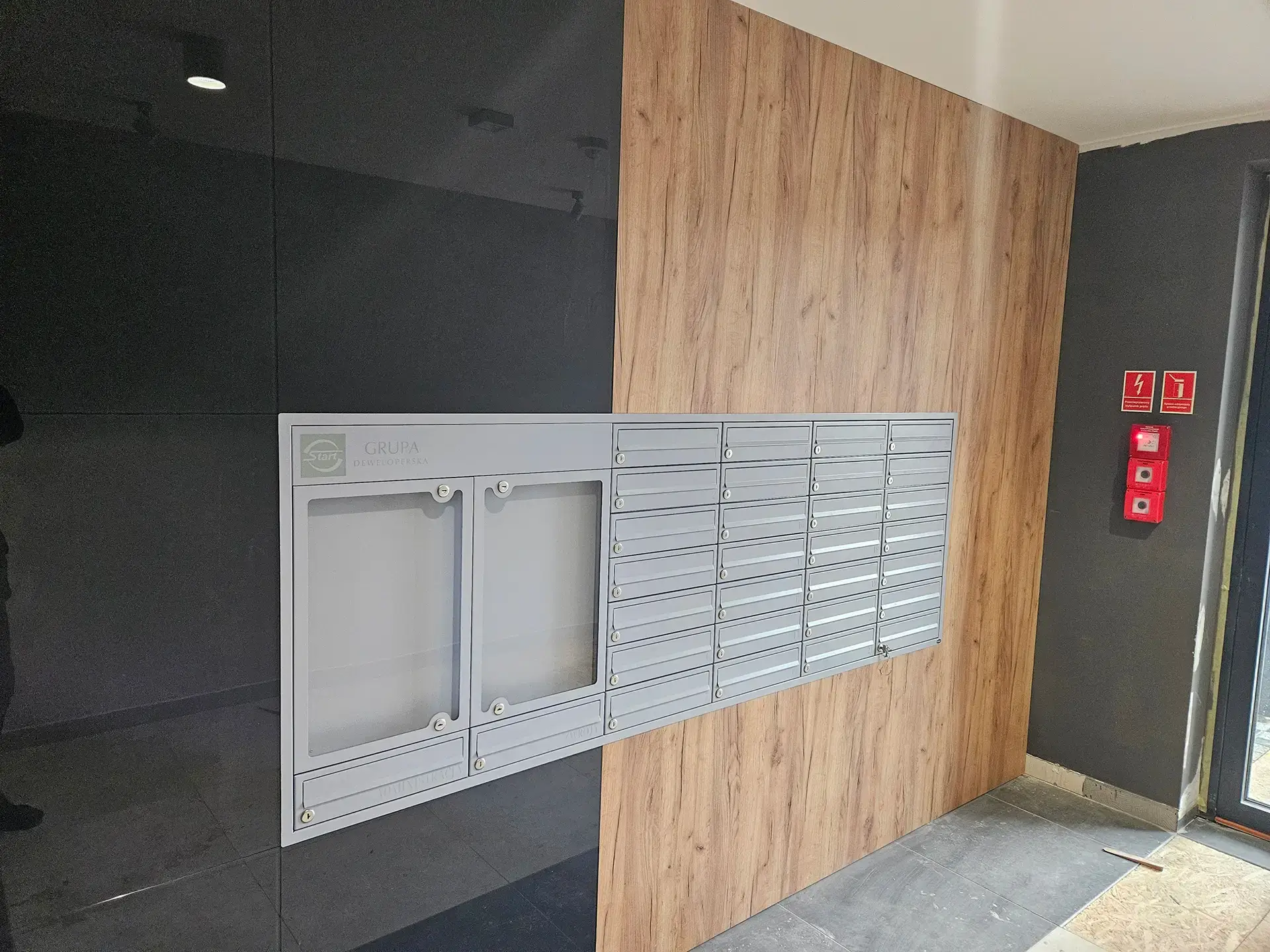 Nowoczesna zabudowa skrzynek pocztowych w biurowcu, łącząca grafitowe panele z elementami drewna.