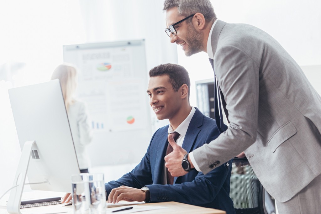 Funcionário trabalhando enquanto gestor faz sinal de jóia com a mão e aprova o trabalho enquanto gráficos de desempenho da empresa aparecem ao fundo