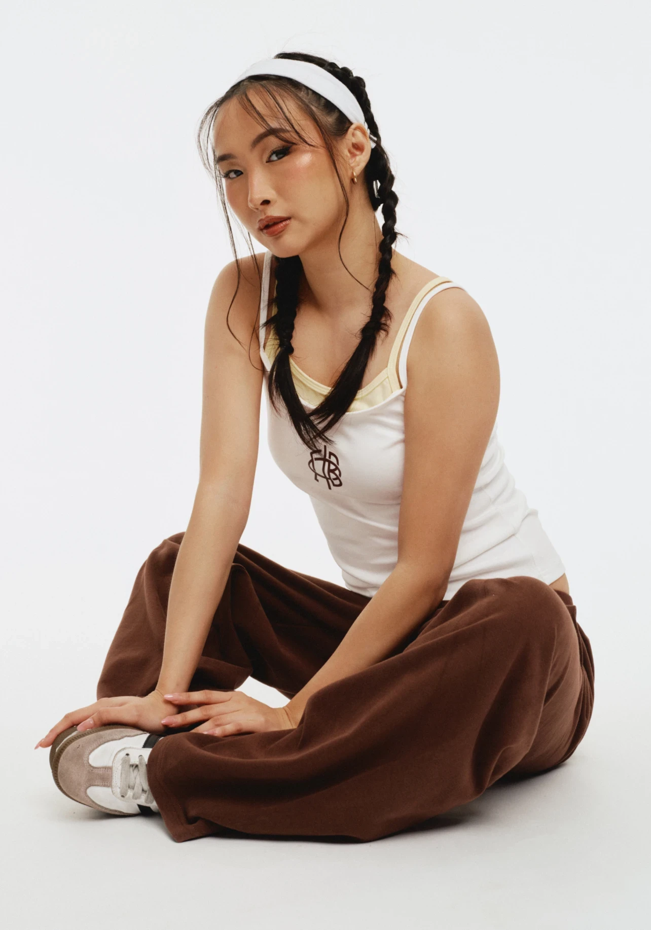 Model seated cross-legged on white backdrop wearing a white tank with yellow trim and a small monogram chest logo, paired with brown sweatpants and a white headband.