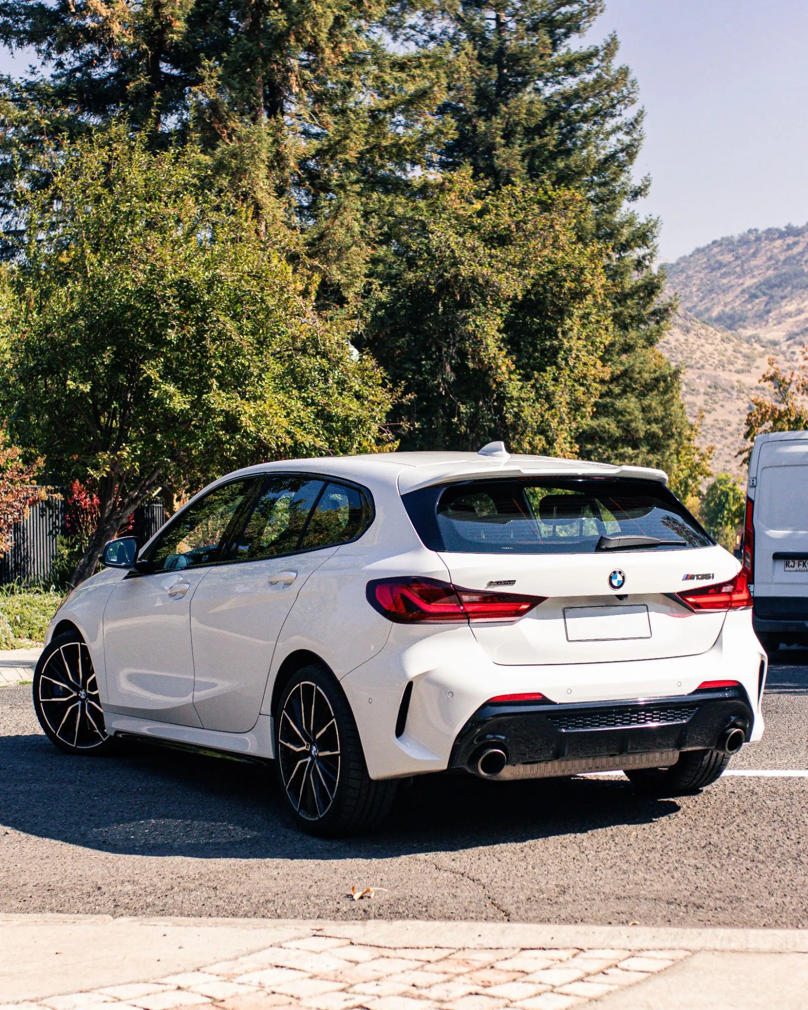 BMW M135i xDrive blanco estacionado en calle arbolada, vista trasera lateral con doble escape