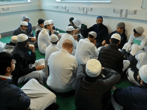 Boys in prayer caps sitting in a circle on a green carpeted floor around a teacher seated against the wall.