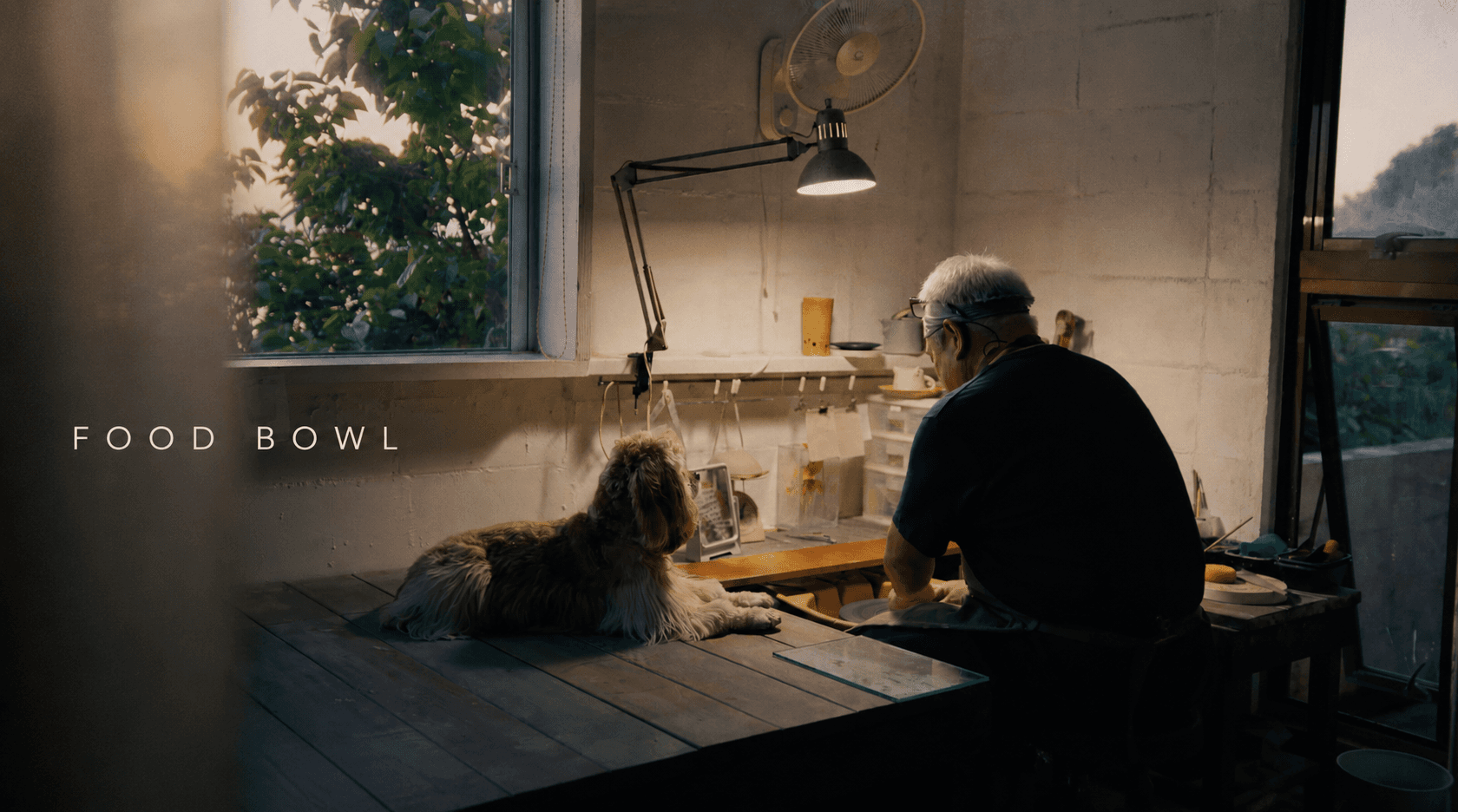 窓辺の作業場で手を動かす職人と、そばに伏せる犬。映像作品「FOOD BOWL」より