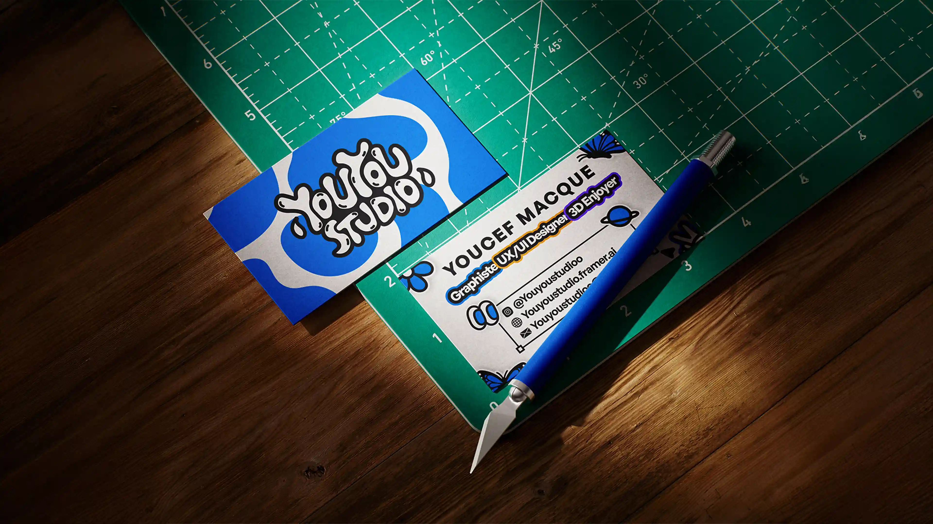 Photo carte de visite recto-verso Youyou Studio pour Youcef Macque - recto avec logo bleu et blanc sur fond argenté, verso avec titre Graphiste UX/UI Designer 3D Enjoyer et coordonnées complètes, disposée sur tapis de découpe vert avec cutter professionnel, ambiance atelier créatif