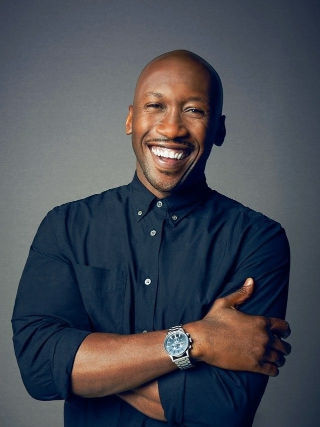 A smiling man with short hair stands with arms crossed, wearing a dark shirt against a neutral background.