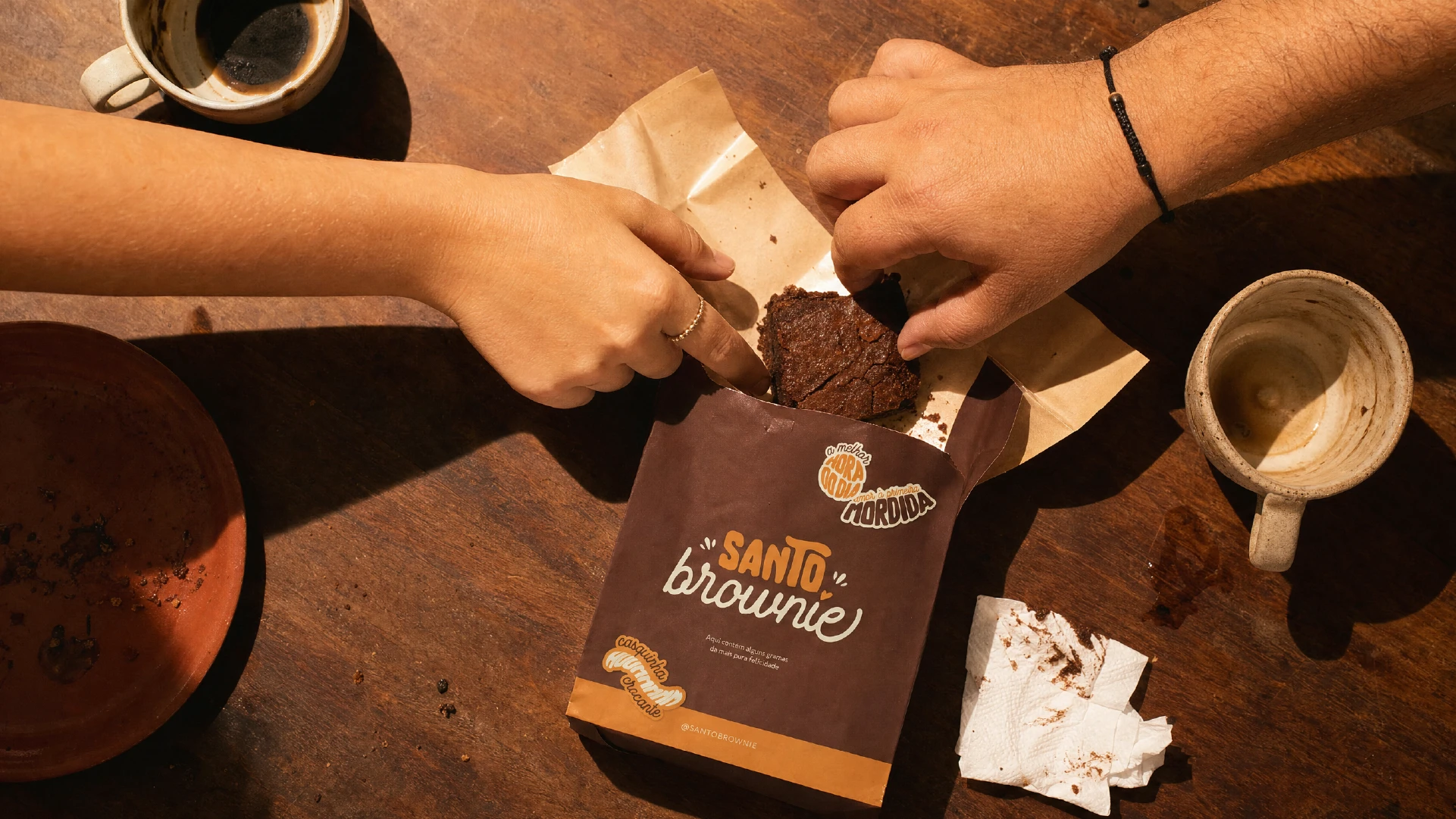 Overhead view of two hands reaching for a brownie piece on parchment paper beside an open Santo Brownie bag, with coffee cups on a wooden table.