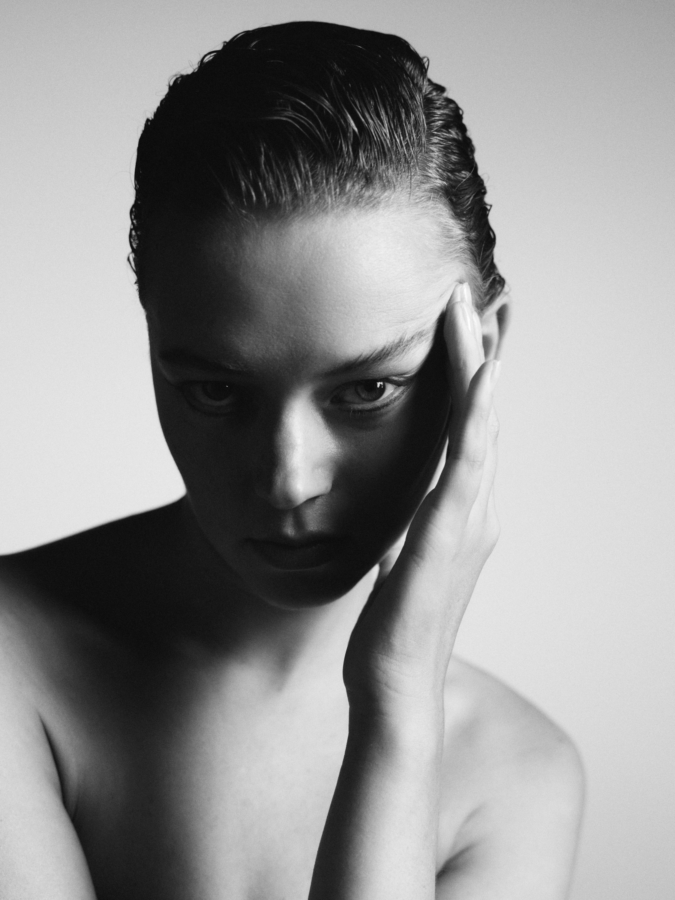 A beauty portrait photo in black and white of a model touching the side of her face. She is looking directly at the camera and her hair is damp and tied back.
