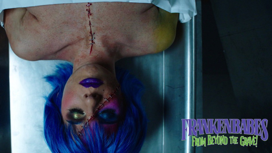 A woman with vibrant blue hair and stitched makeup lies on her back, appearing lifeless, with dramatic lighting casting shadows on her face.