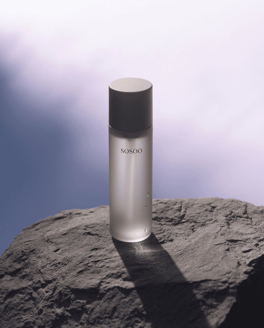 A frosted bottle of SOSOO skincare stands on a rough rock against a soft, blurred sky background. The scene evokes tranquility and elegance.