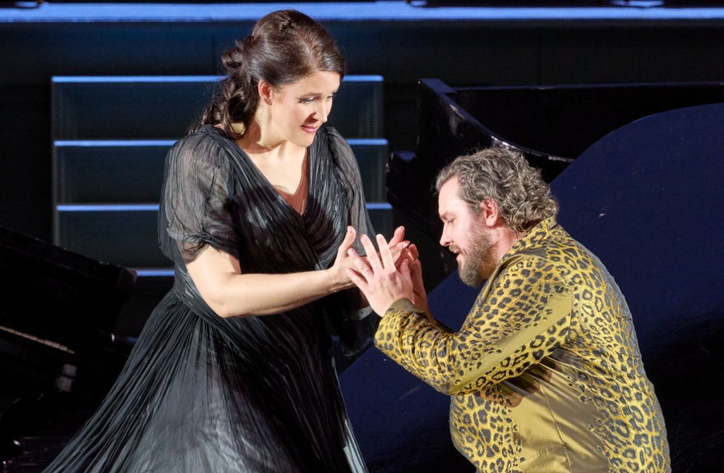 A woman in black dress holds the hands of a man in a golden coat kneeling before her