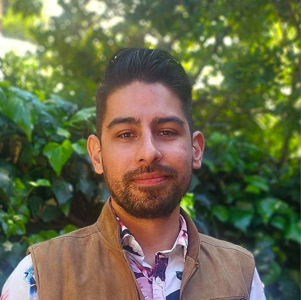Retrato de uma pessoa com cabelo curto e escuro e uma barba bem aparada, vestindo uma camisa estampada e um colete bege, olhando para a câmera com um sorriso suave, em pé ao ar livre com folhagem verde e luz do sol ao fundo.