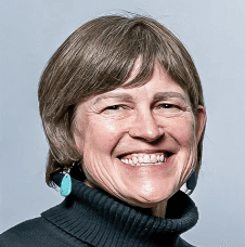 A smiling woman with short brown hair and a dark turtleneck sweater, wearing turquoise teardrop earrings, representing.