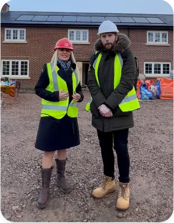 Two people wearing high-visibility vests and hard hats standing on a construction site in front of a new brick house development