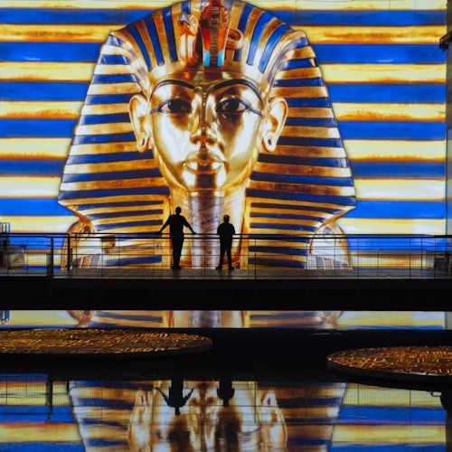 บุคคลสองคนในเงายืนอยู่บนทางเดินหน้าภาพแสดงหน้ากากของตุทันคาเมนที่มีแสงสว่างสดใส พร้อมเงาสะท้อนในน้ำ