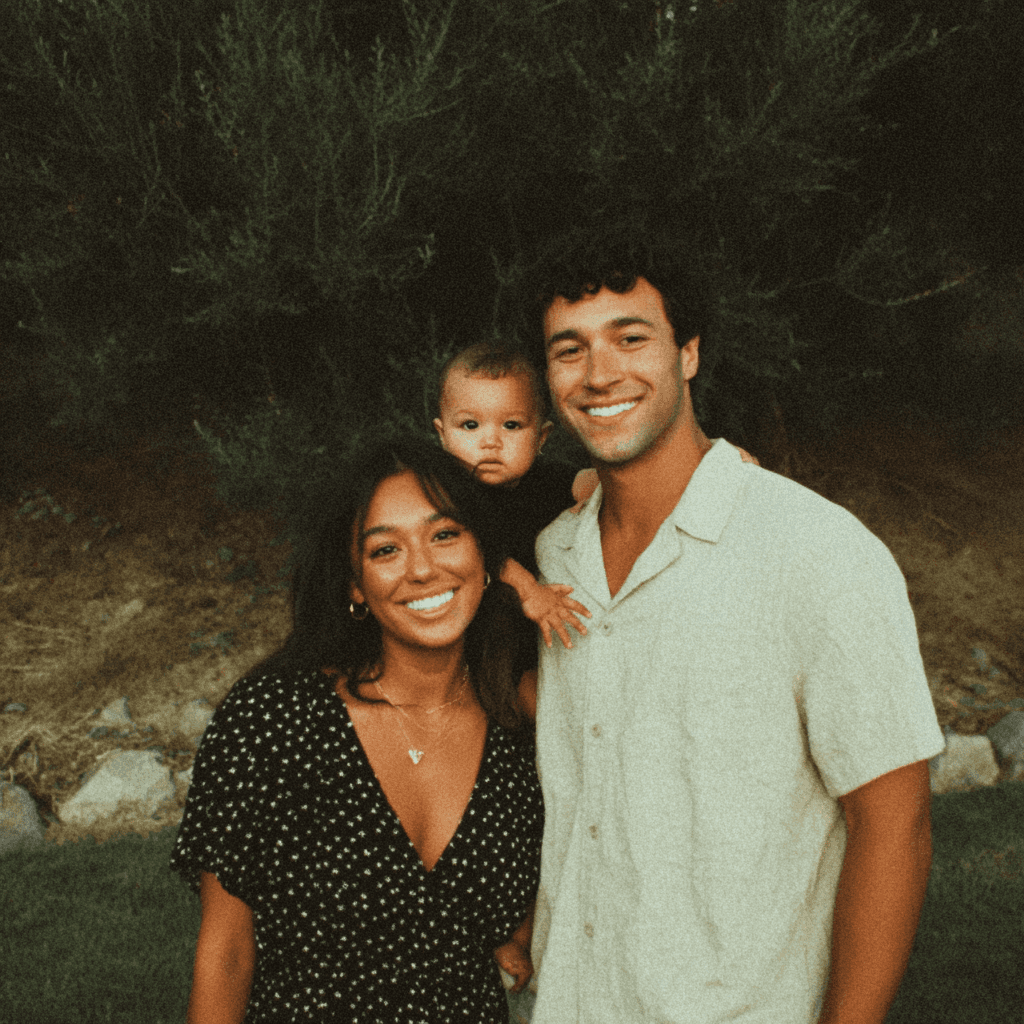 A joyful family moment is captured as a man and woman, both smiling, playfully embrace while a happy baby clings onto the man's shoulders, set against a lush, green outdoor backdrop.