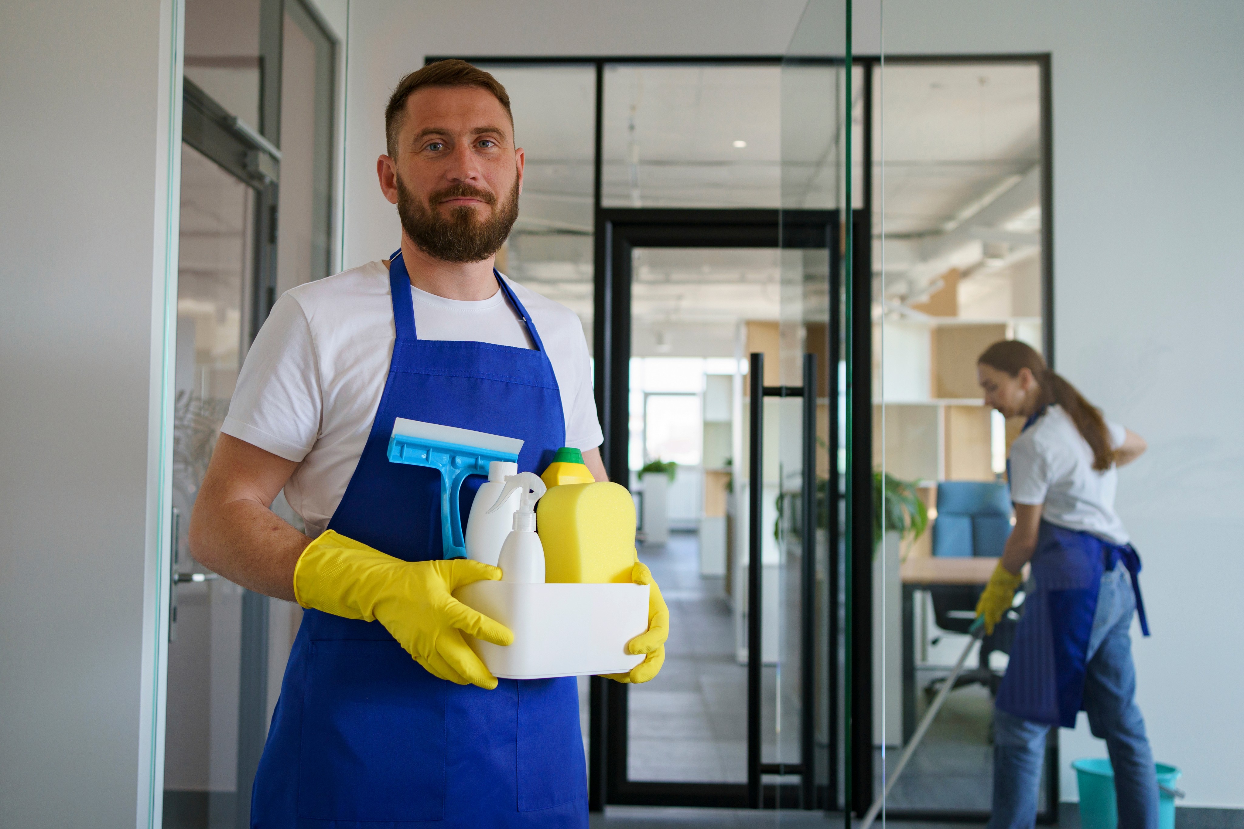 Portrait of a male professional cleaner holding a bucket of cleaning supplies and detergents, standing in a blurred hallway.