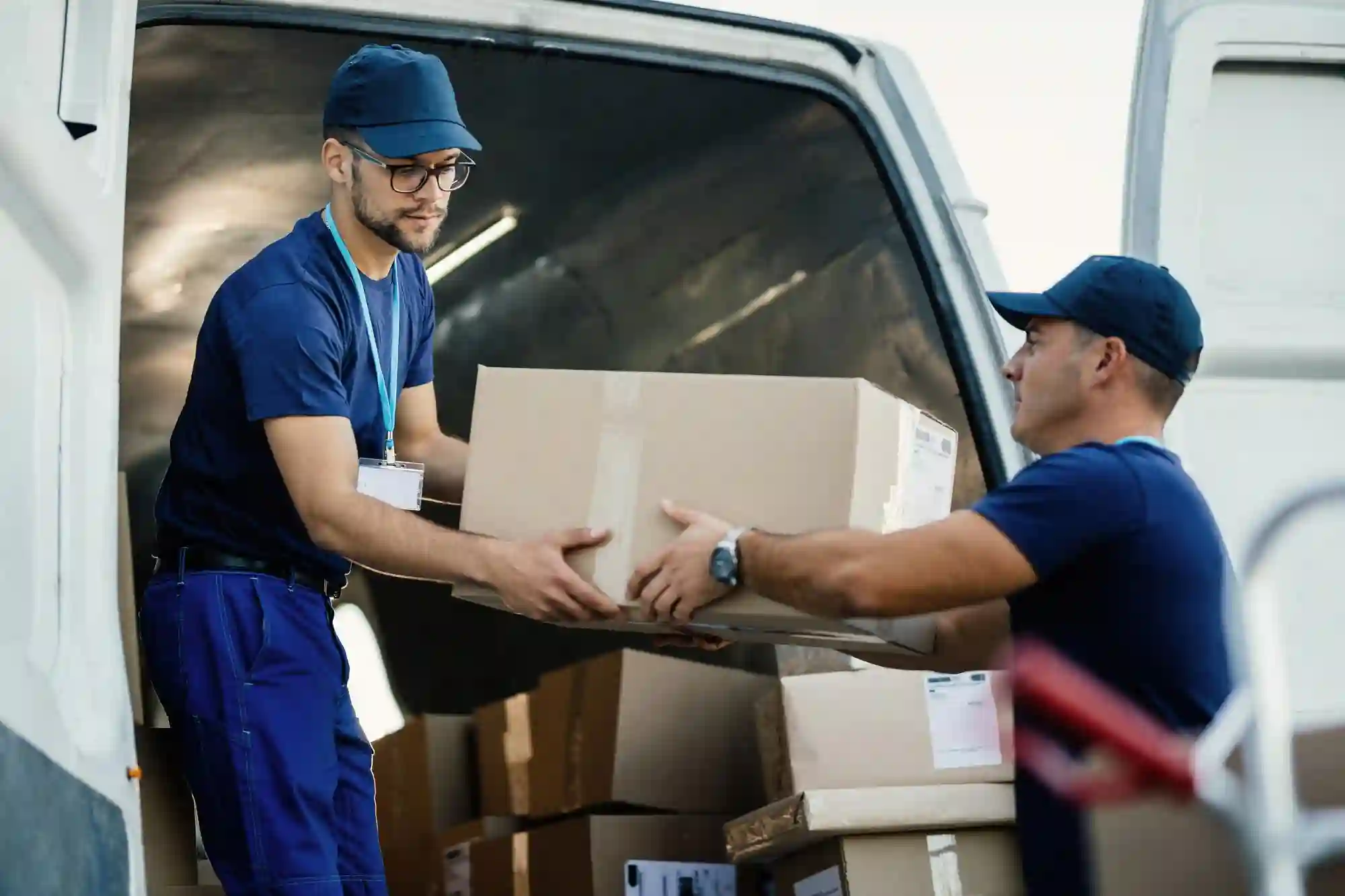 Two delivery workers in blue uniforms unloading packages from the back of a white delivery truck.