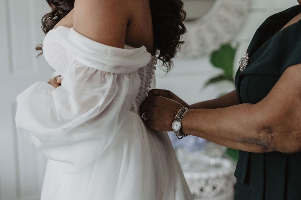 Bridal attendant fastening the bride’s off-the-shoulder gown.