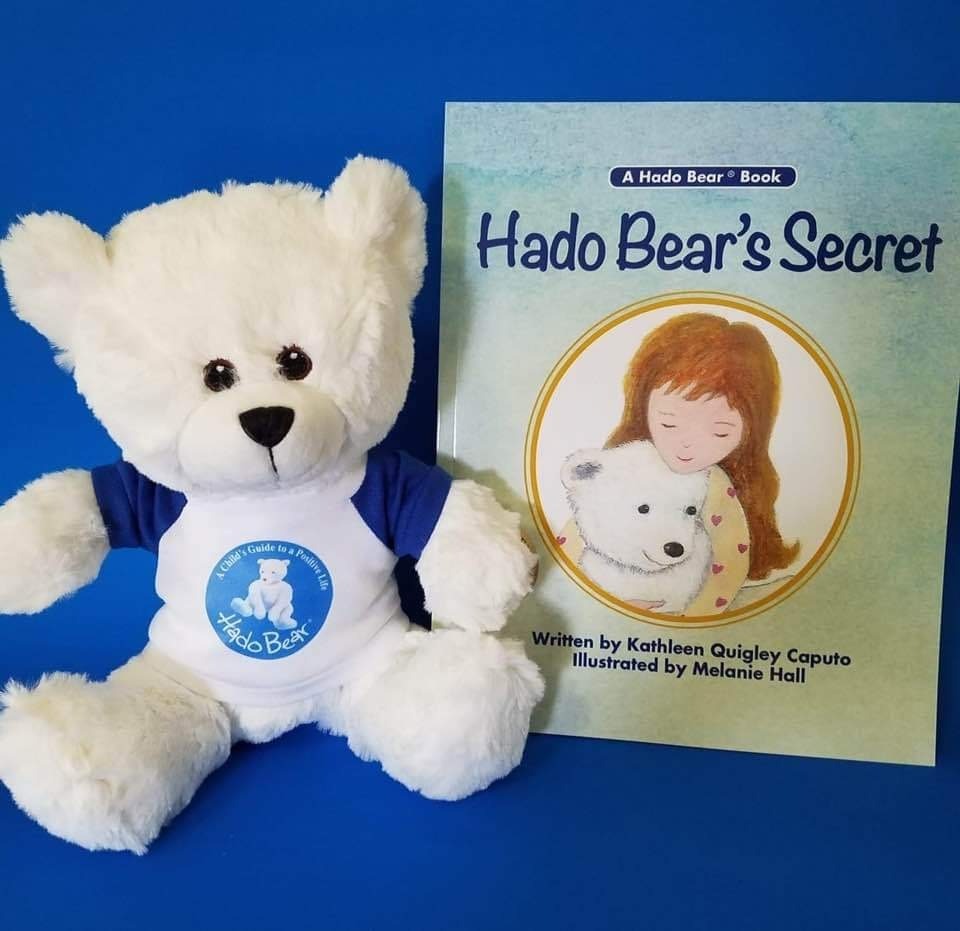 A white teddy bear wearing a blue and white shirt sits beside a children’s book titled "Hado Bear's Secret" against a blue background. The book cover features an illustration of a young girl hugging a white bear, conveying warmth and affection.