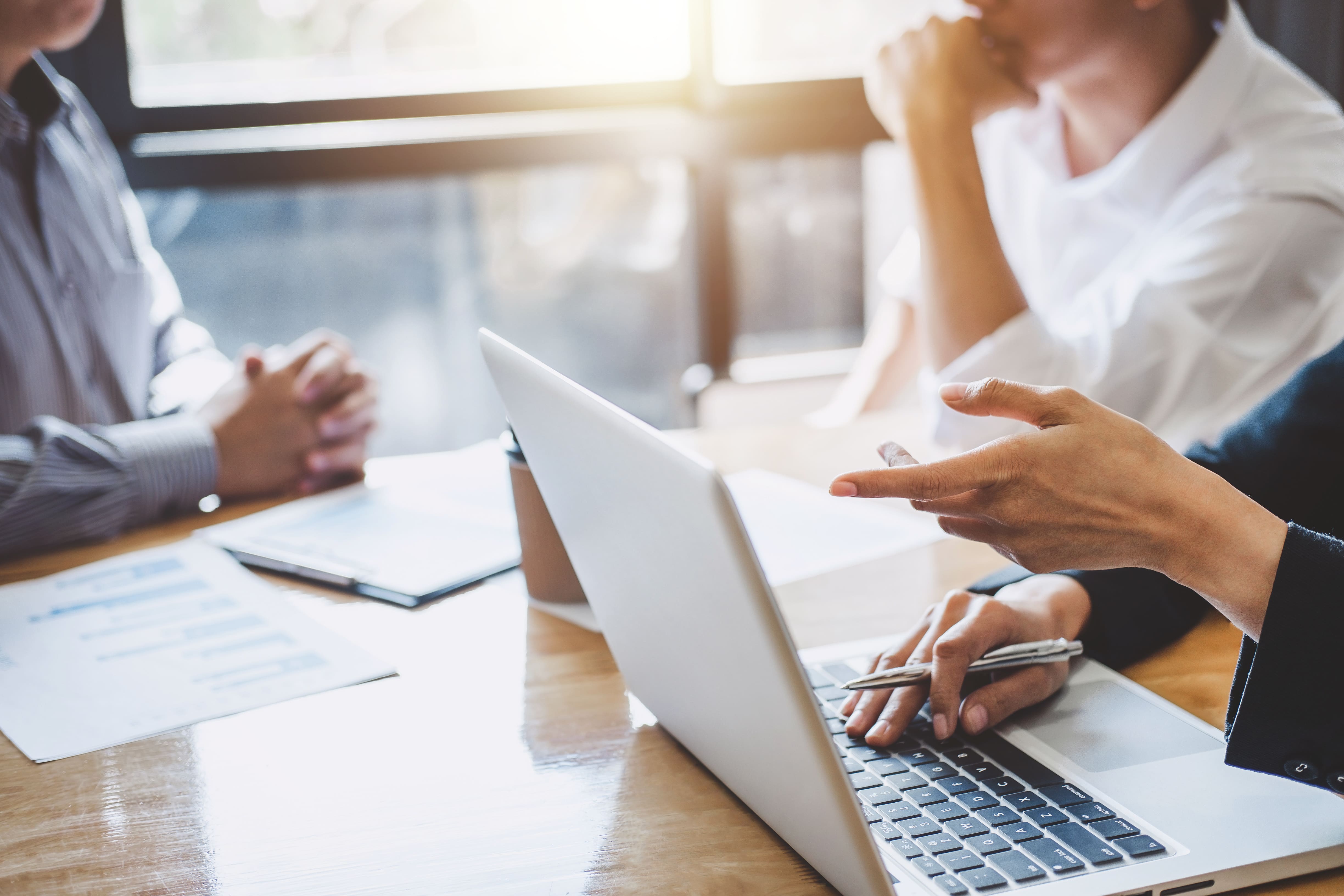A group of people sits around a table in a bright office, engaging in a discussion with laptops and documents, highlighting a collaborative work environment.