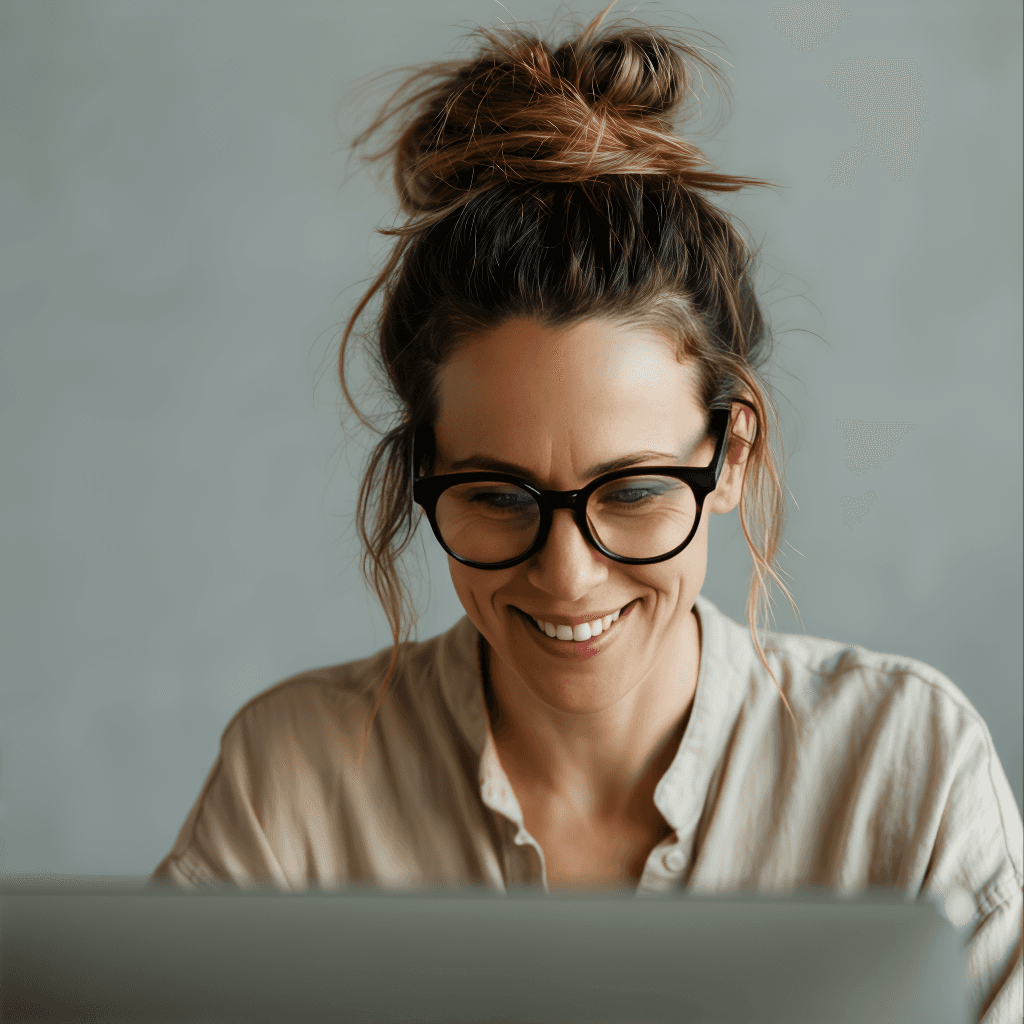 Woman with glasses smiling while working on a laptop. She has a messy bun hairstyle and tan blouse.
