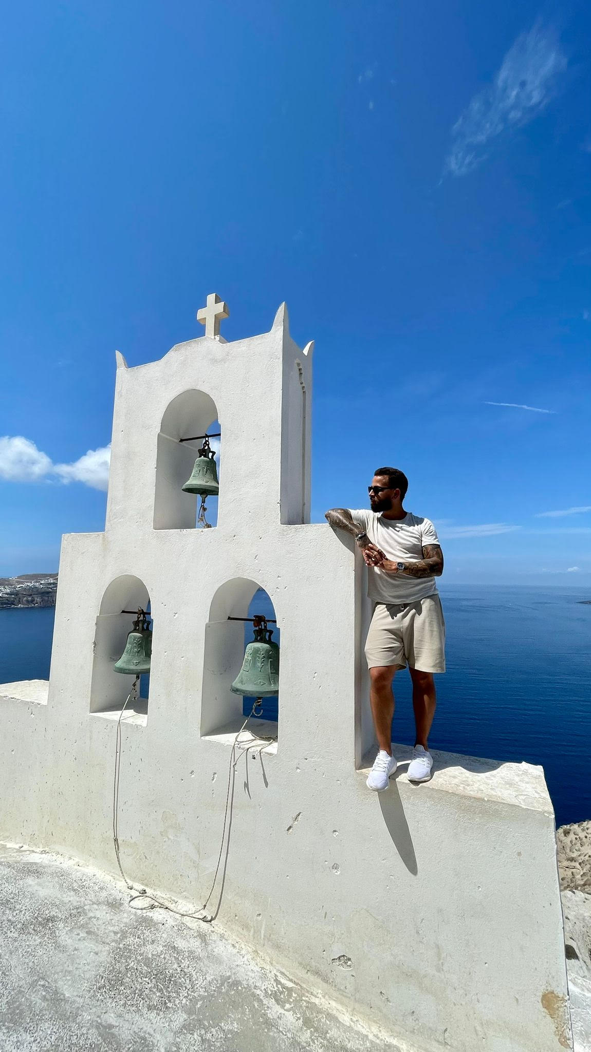 David in a white t-shirt and shorts leans against a white bell tower with three bells, overlooking the ocean. He is wearing sunglasses and looking to the side, with a clear blue sky in the background.