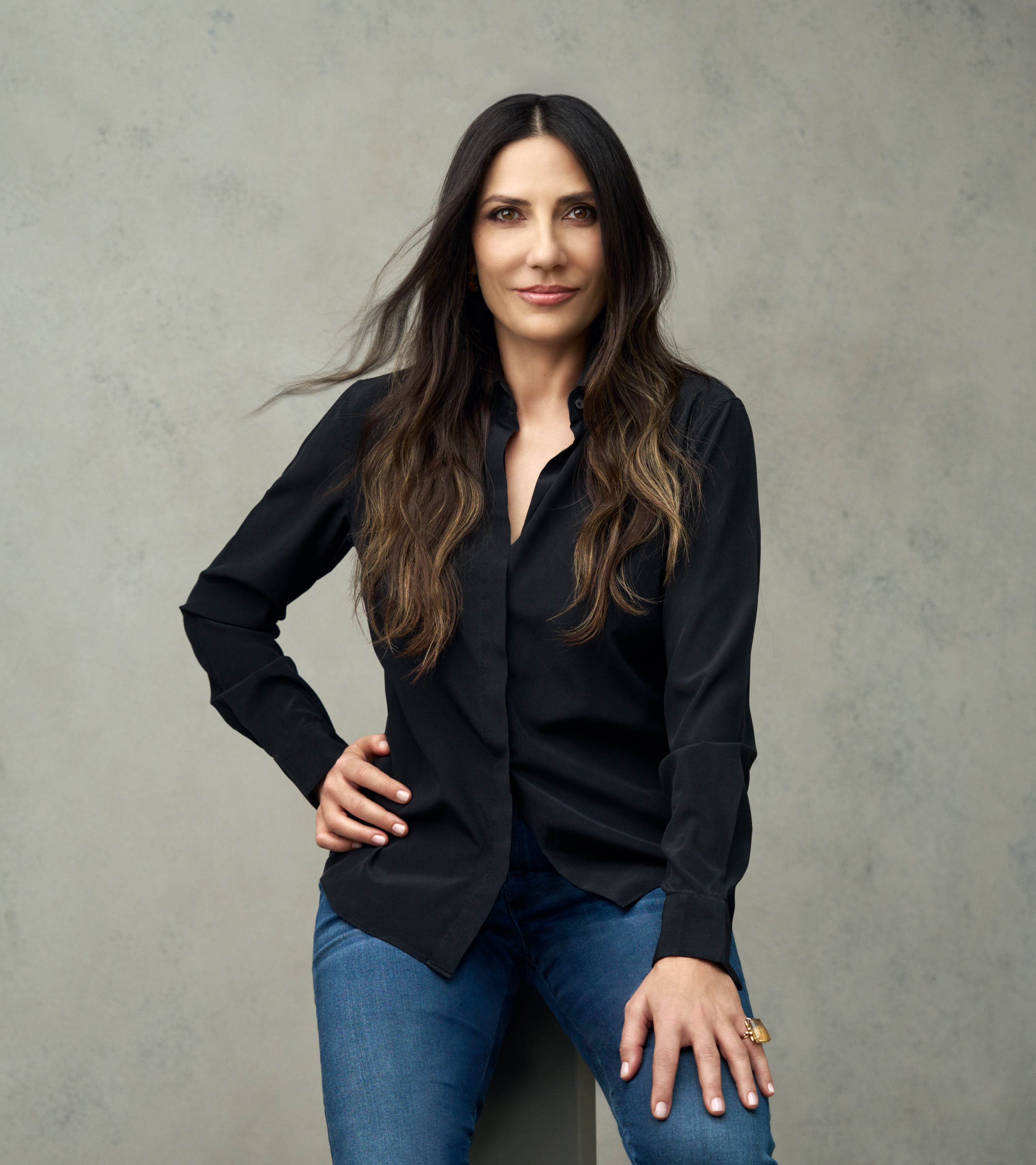 A young person with curly hair smiles at the camera while sitting in front of a laptop against a dark background.