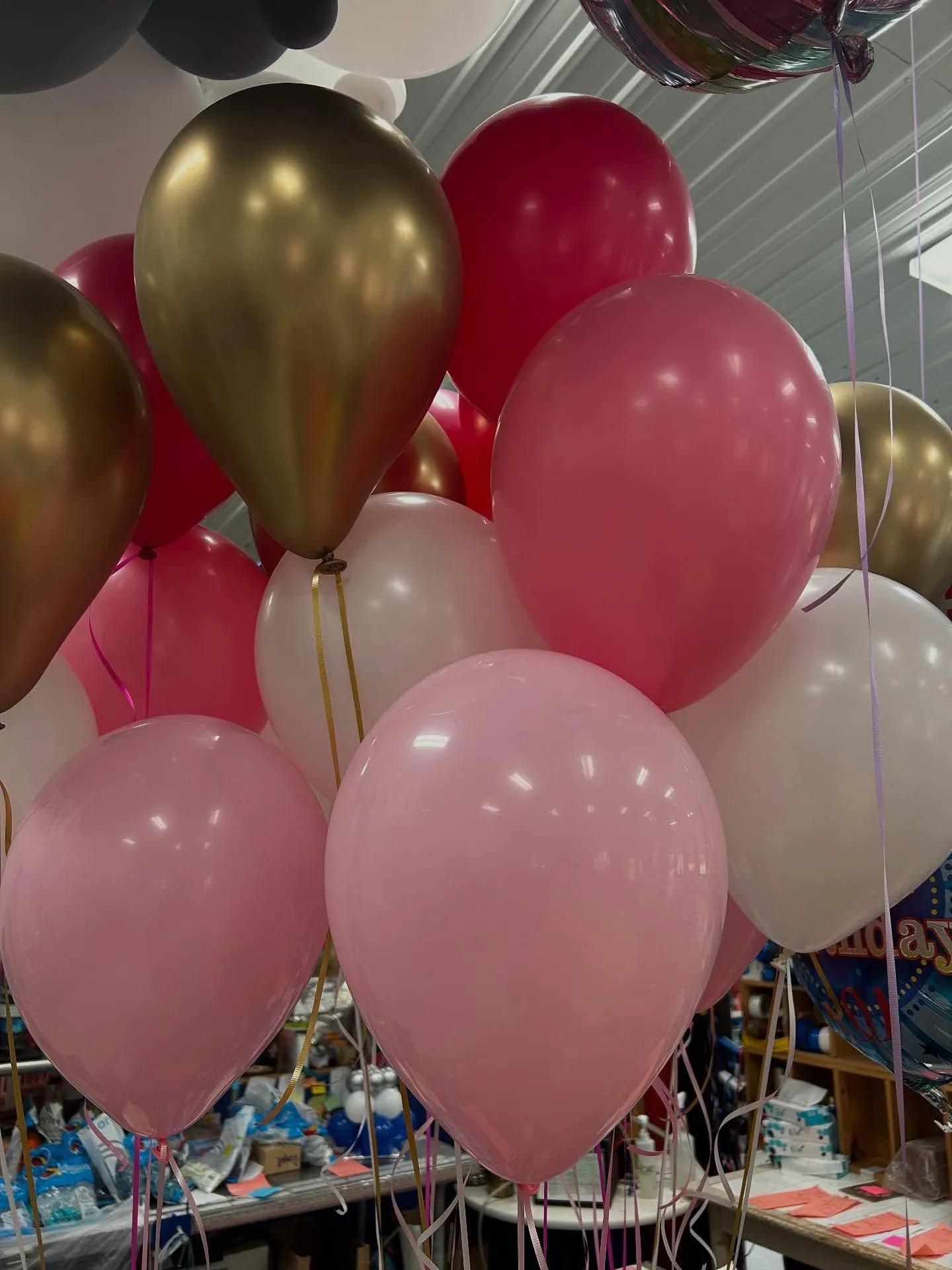 A festive assortment of gold, pink, and white helium balloons float under a vaulted ceiling, creating a joyful celebration atmosphere with vibrant colors.