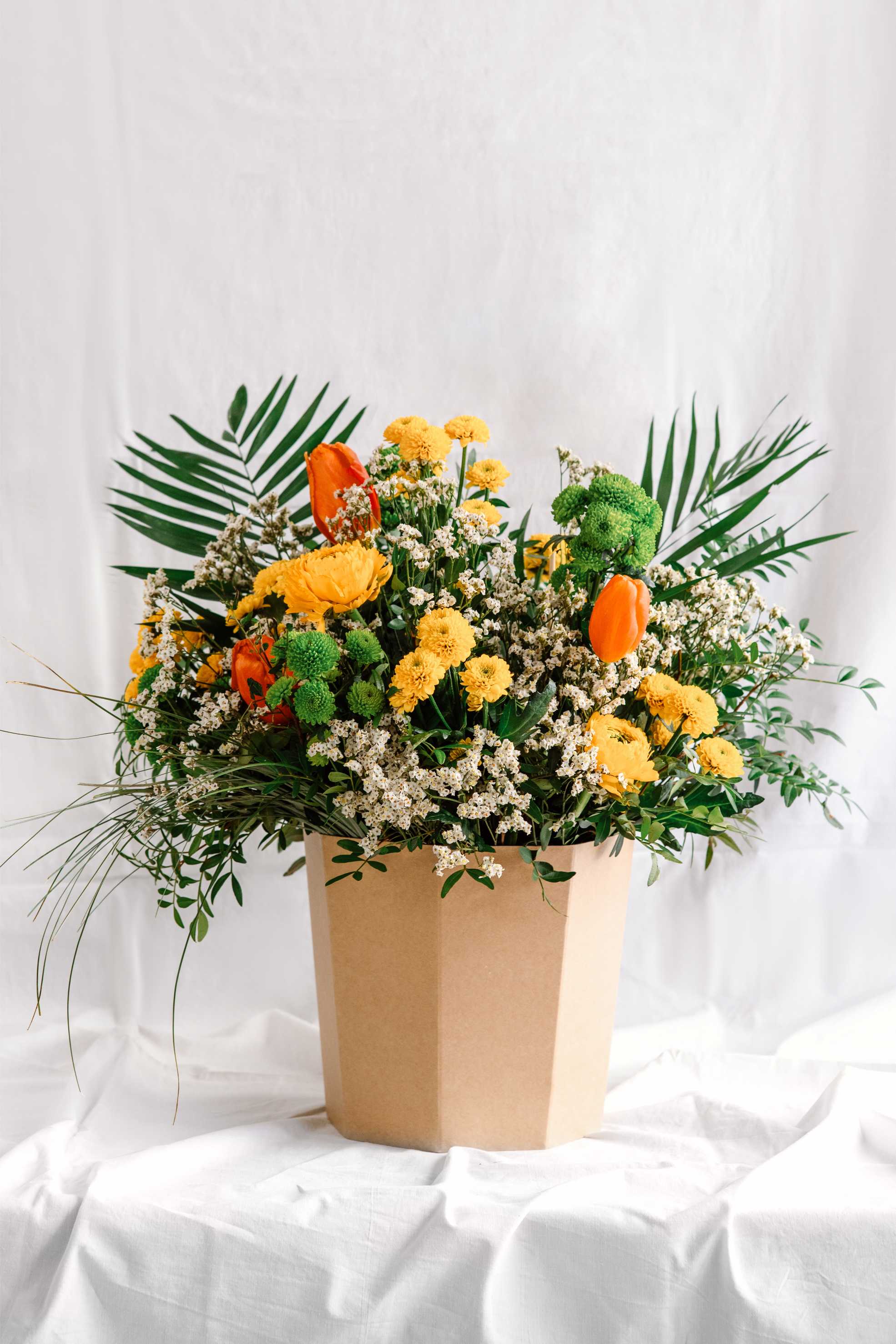 Arranjament floral en caixa de cartró kraft amb flors de temporada grogues, taronges i verdes, destacant crisantems, ranuncles, tulipes i verds decoratius com palmes i flor d’arròs.