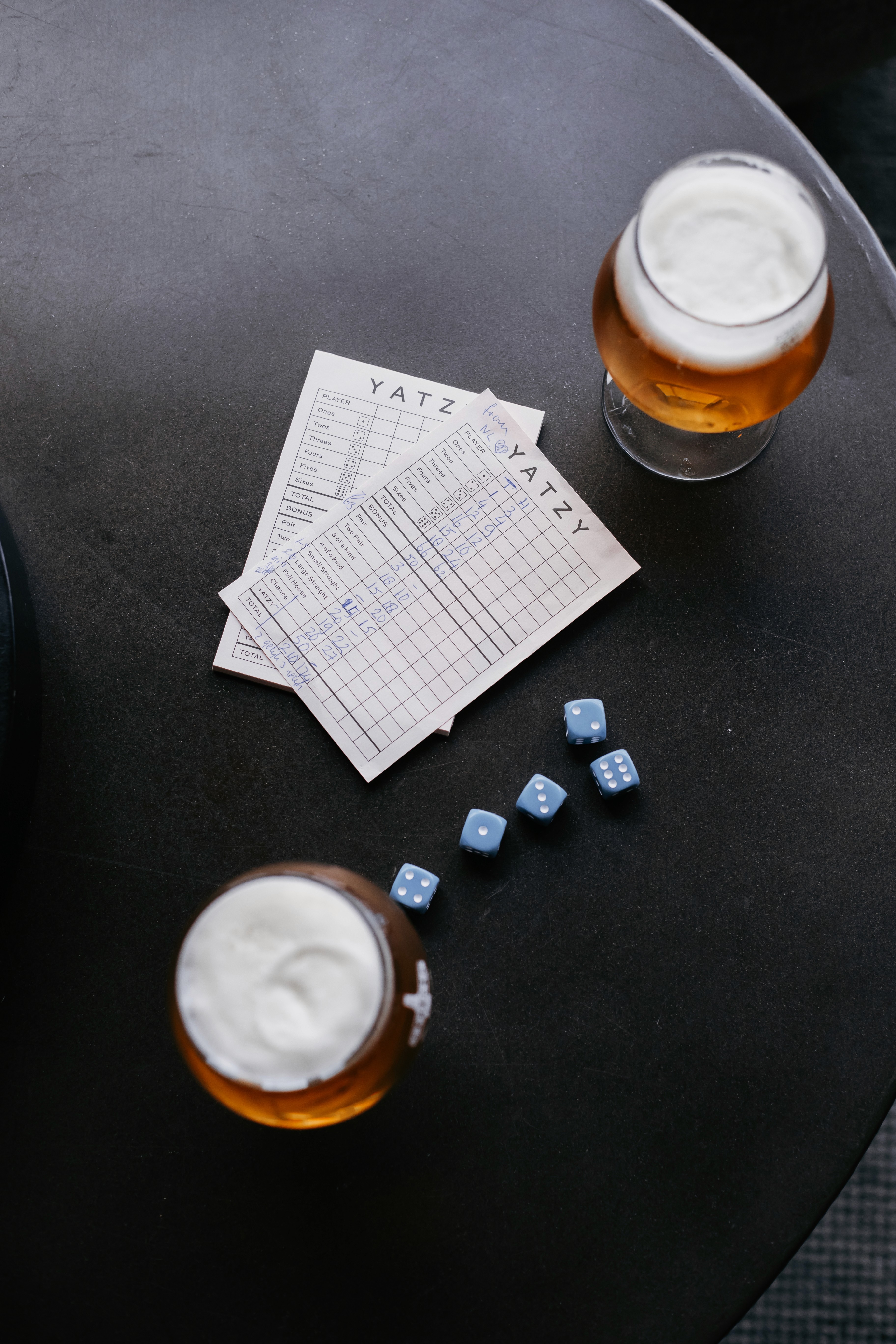 Yatzy scorecards and blue dice beside two glasses of beer on a dark table