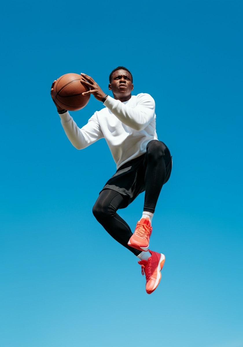 Dynamic shot of a basketball player in mid-air, wearing red sneakers and a white shirt, set against a clear blue sky.