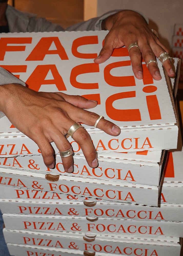 Men wearing silver rings with a stack of pizza and focaccia boxes from restaurant Facci with hands reaching for one. 