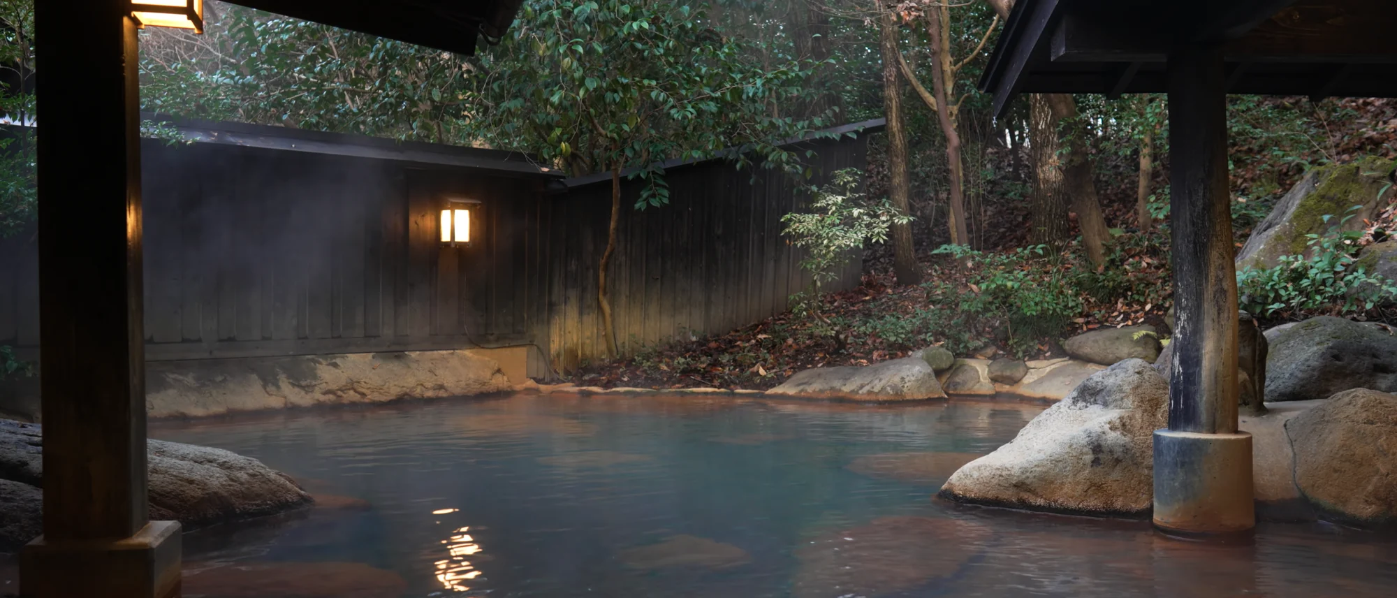 A wide open-air hot spring bath of pale blue-green water enclosed by boulders and forest