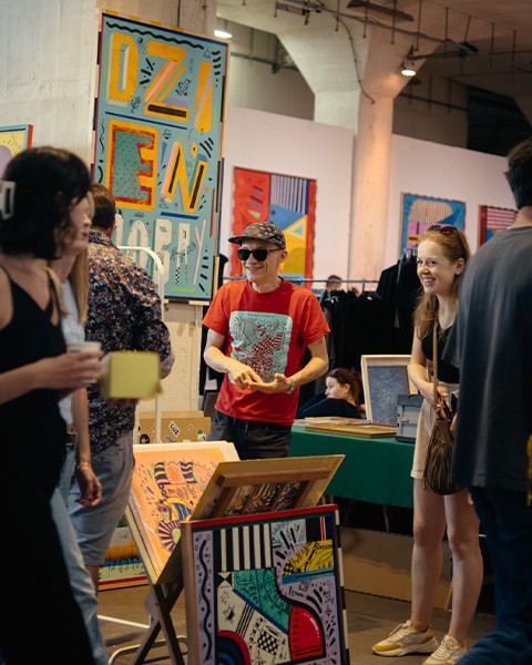 Zdjęcie przedstawiające artystę na stanowisku targowym, pośród swoich prac i grupy ludzi z nim rozmawiających.
