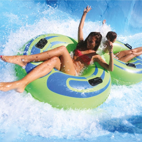 Two people in swimsuits enjoy a water slide, each sitting in green and blue inflatable tubes, splashing into the water.