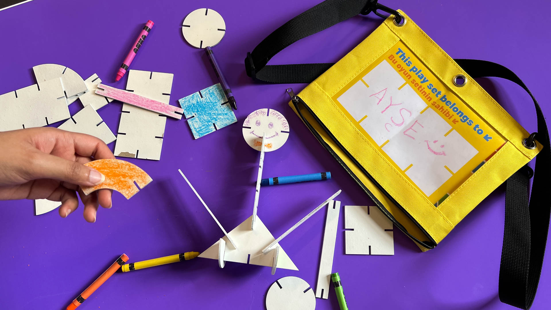 A yellow fabric pouch with a black strap lies on a purple surface. The pouch has a white label with the words “This play set belongs to AYSE” and a child’s drawing of a smiling face. Scattered around the pouch are various craft supplies: wooden shapes, colored pencils, and small circular cutouts. A hand holds a piece of orange-red material near the pouch.