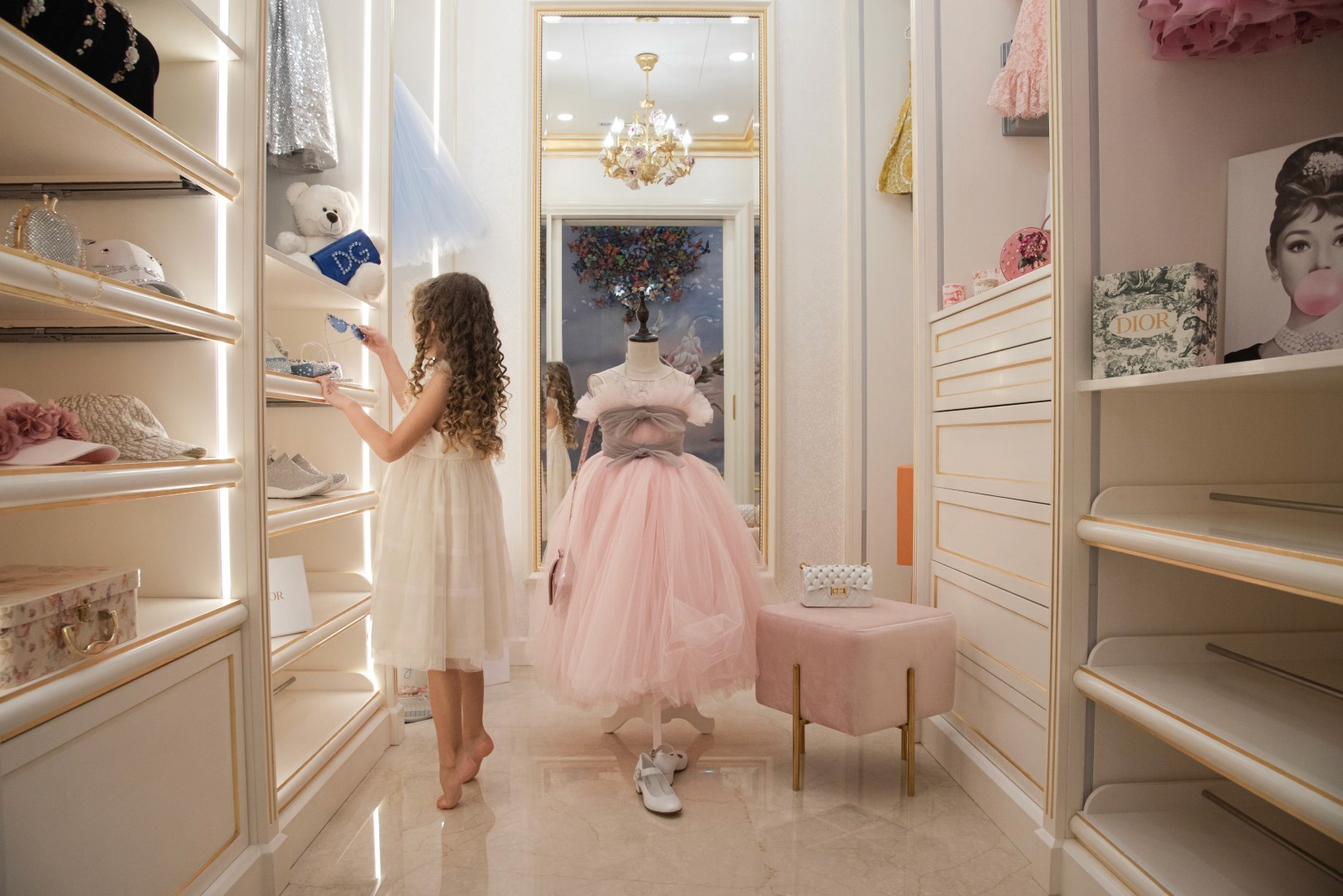A young girl in a white dress is selecting items in a luxurious walk-in closet, featuring elegant pastel décor, a soft pink tulle dress on a mannequin, and neatly arranged shelves with assorted fashion accessories.