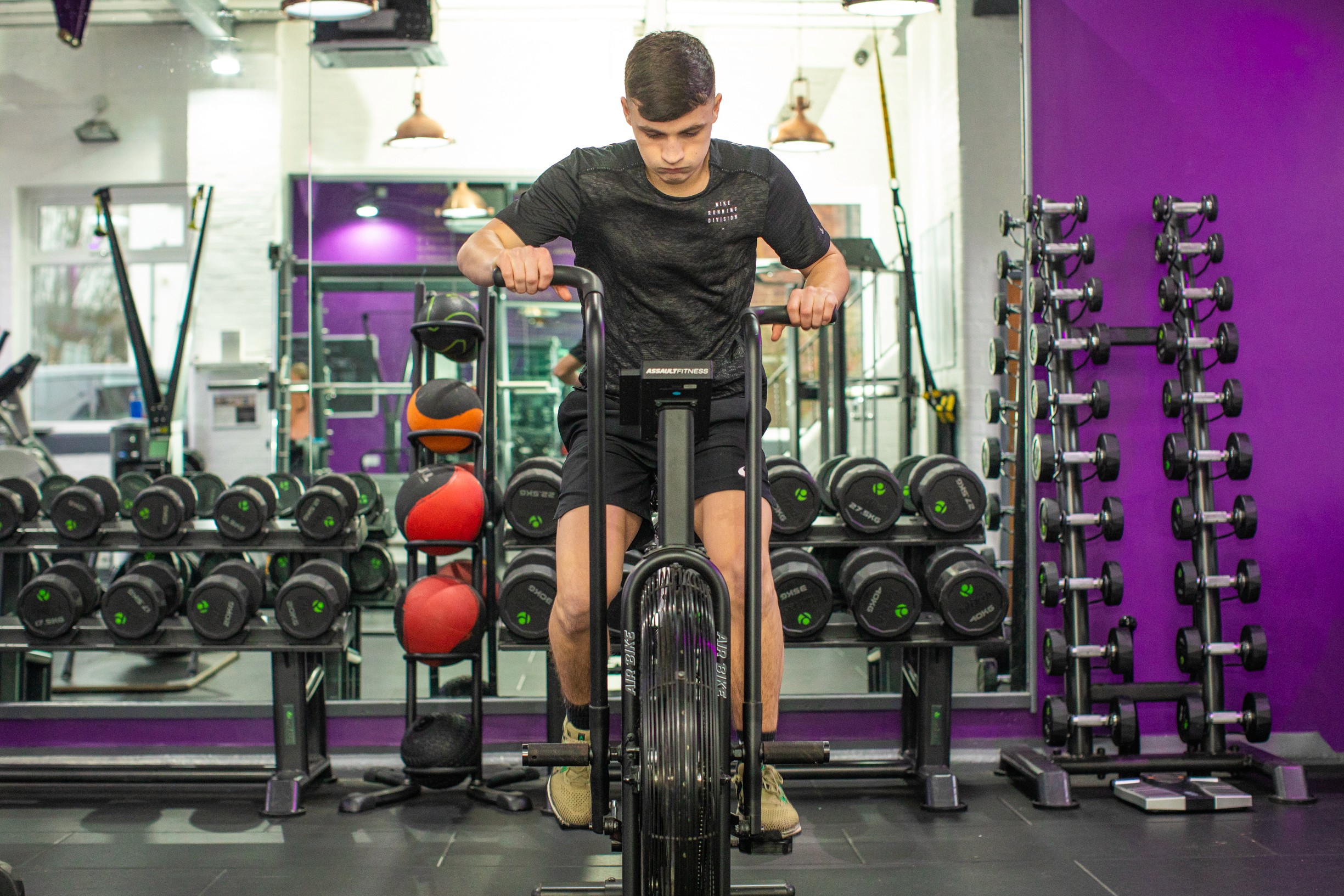 A person uses an air bike in a gym, surrounded by dumbbells, medicine balls, and other exercise equipment against a purple wall.