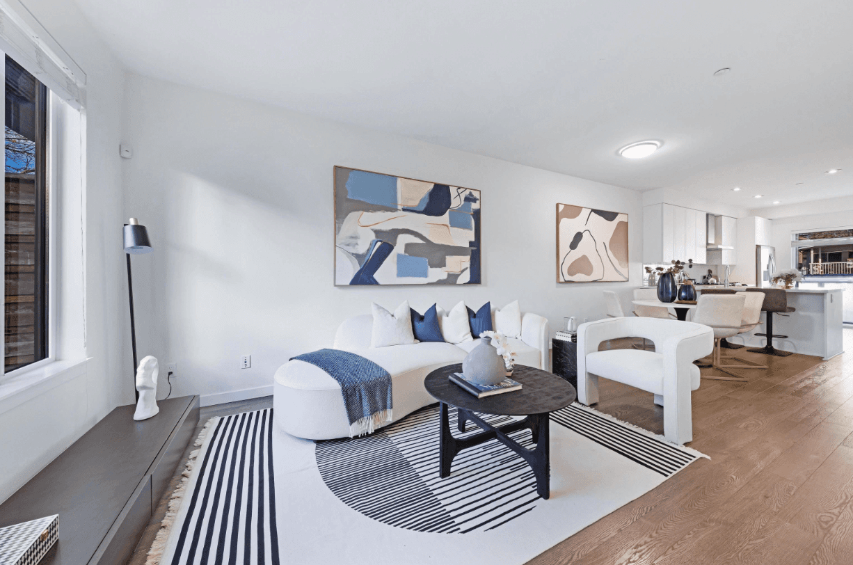 Bright, modern living room with white walls, blue accents, and a cozy seating area. Artwork and wooden flooring visible.