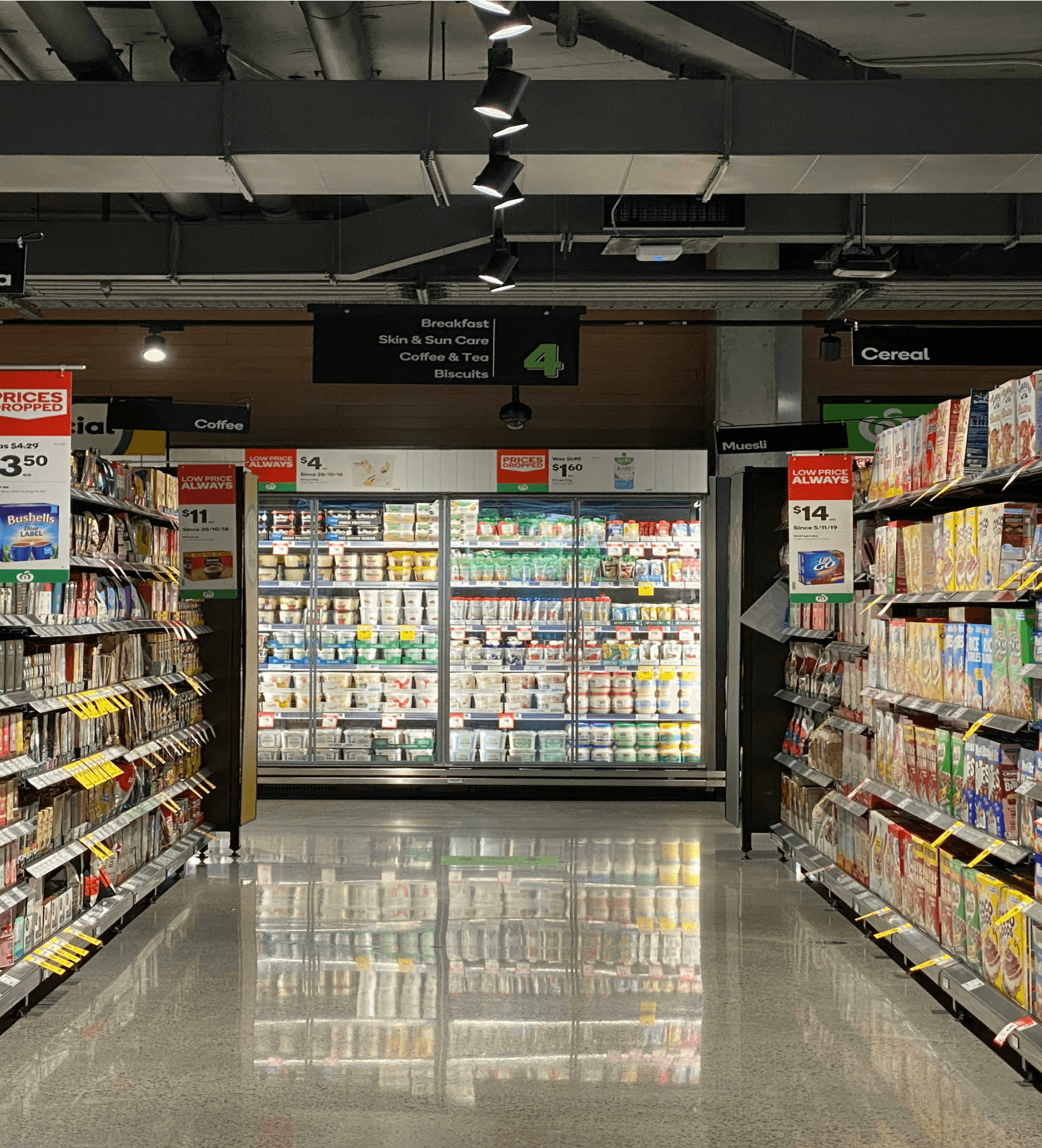 A grocery store aisle with shelves stocked with products, leading to a refrigerated section at the end.