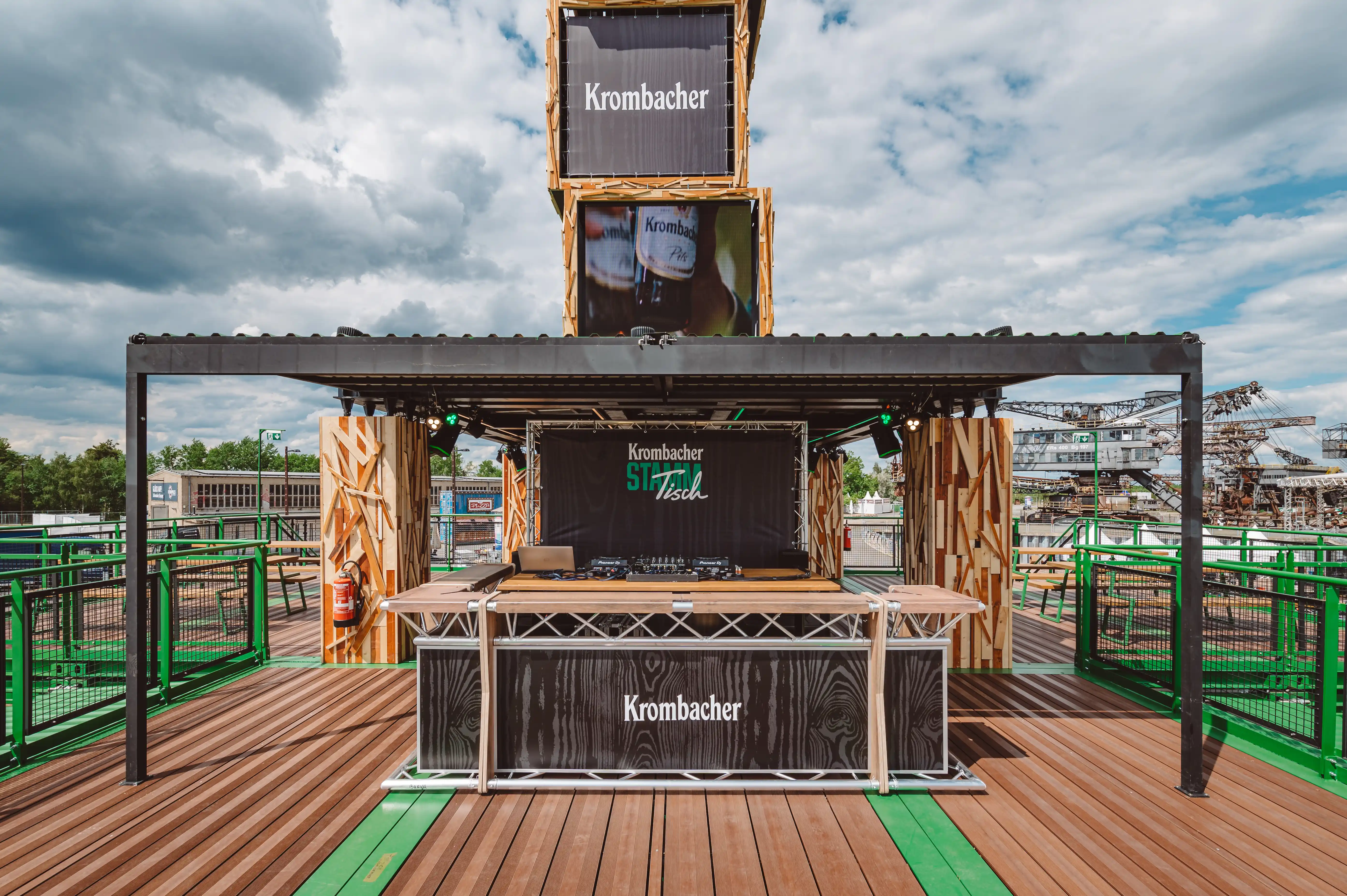 DJ bar on the KROMBACHER roof terrace with a black pergola, wood-clad columns, branded counter and the crate tower with screens above.