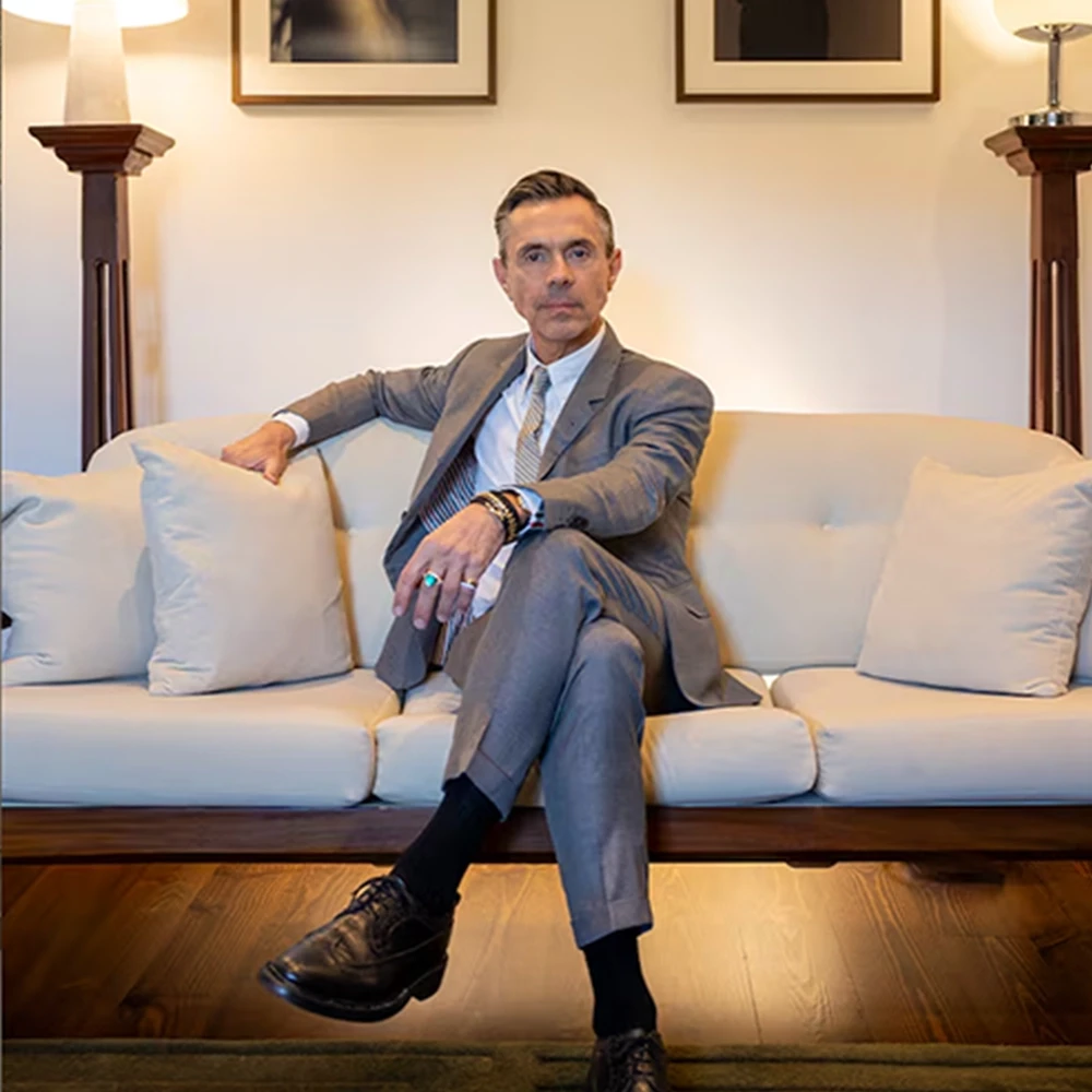 Portrait of British master jeweller Shaun Leane dressed in a tailored grey suit, seated cross-legged on a cream sofa inside a warmly lit, elegant atelier interior setting.