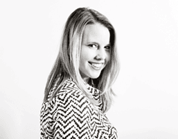 A black and white portrait of a woman with shoulder-length blonde hair, smiling and looking over her shoulder. She wears a black and white chevron-patterned top. The background is plain white.